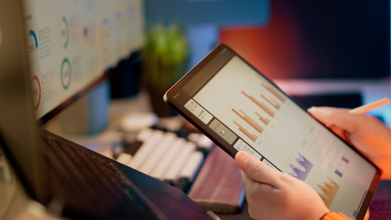 Hands holding a tablet showing performance charts, with a laptop and screens behind