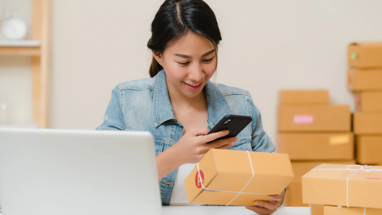 Small business owner checking her phone beside stacked parcel boxes