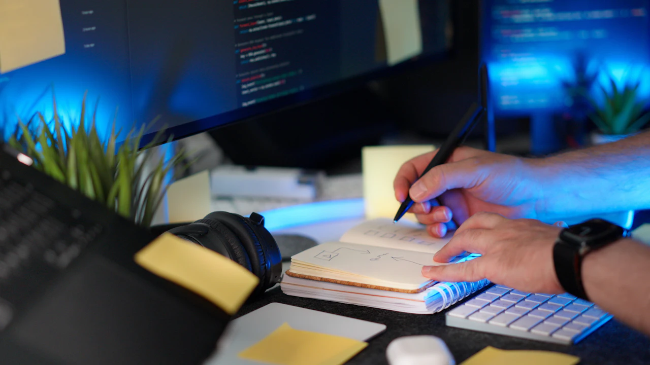 Hands sketching a page flow in a notebook beside monitors showing code